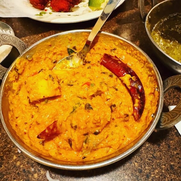 A close-up of a Maa Kitchen Indian curry dish with paneer and a red chili in a metal bowl.